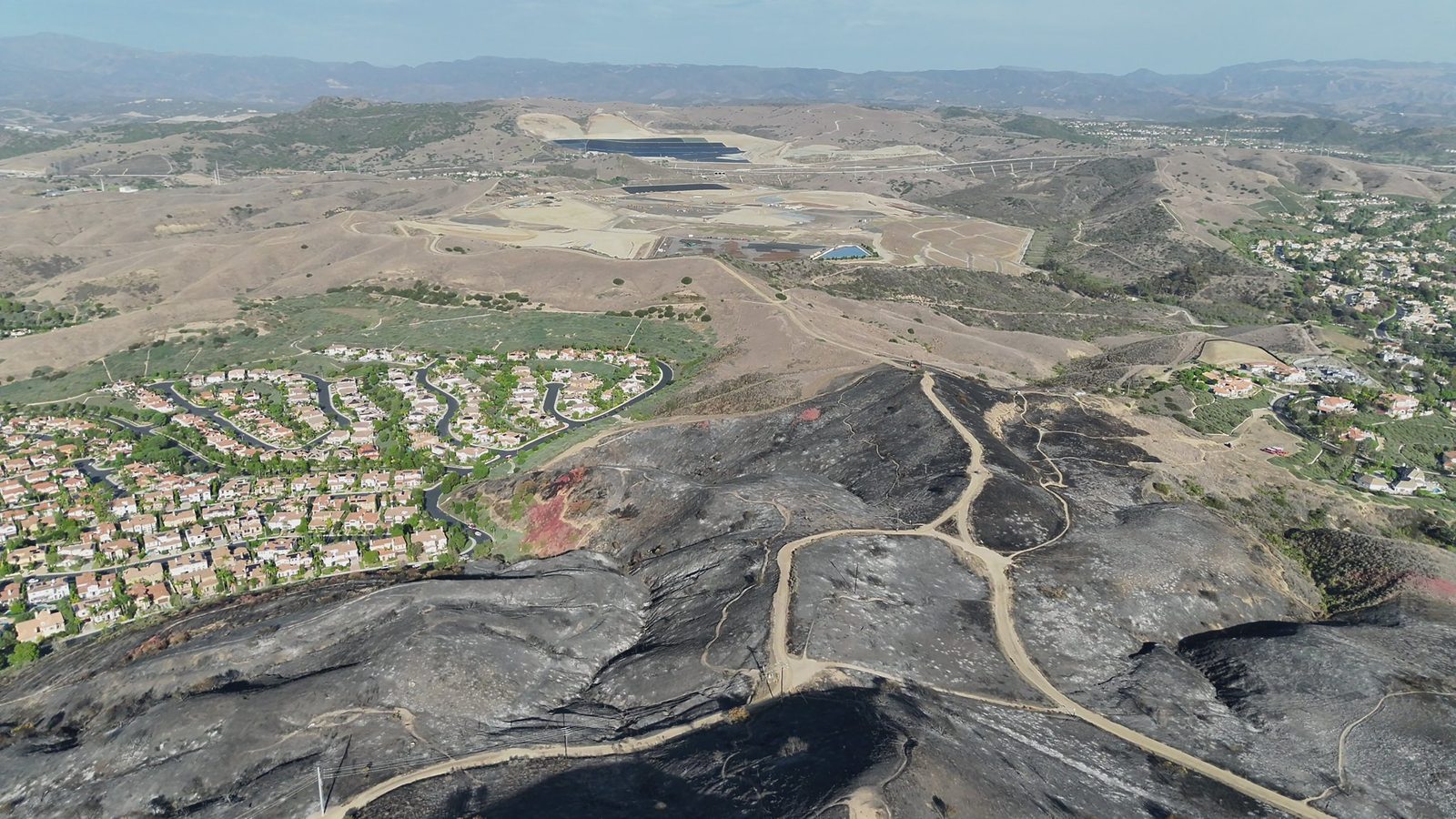 An aerial view shows hillsides scorched by the Norte Fire beside residential neighborhoods in San Clemente, with lines of pink retardant still visible. The fire held at 570 acres on Sunday as containment reached 60 percent.